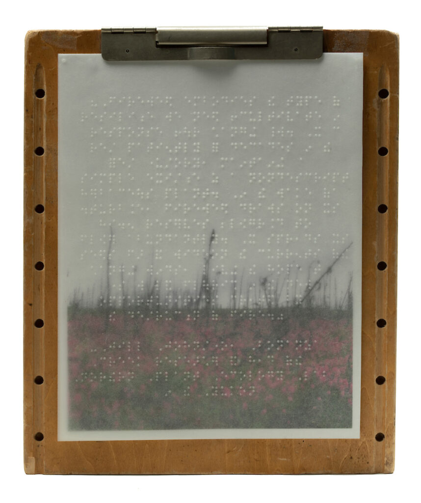 This is a wooden Braille slate that looks like a clipboard with a thin waxed sheet with Braille written on it. A photo transparency rests behind the Braille and is attached by the clipboard mechanism as well as taped in place